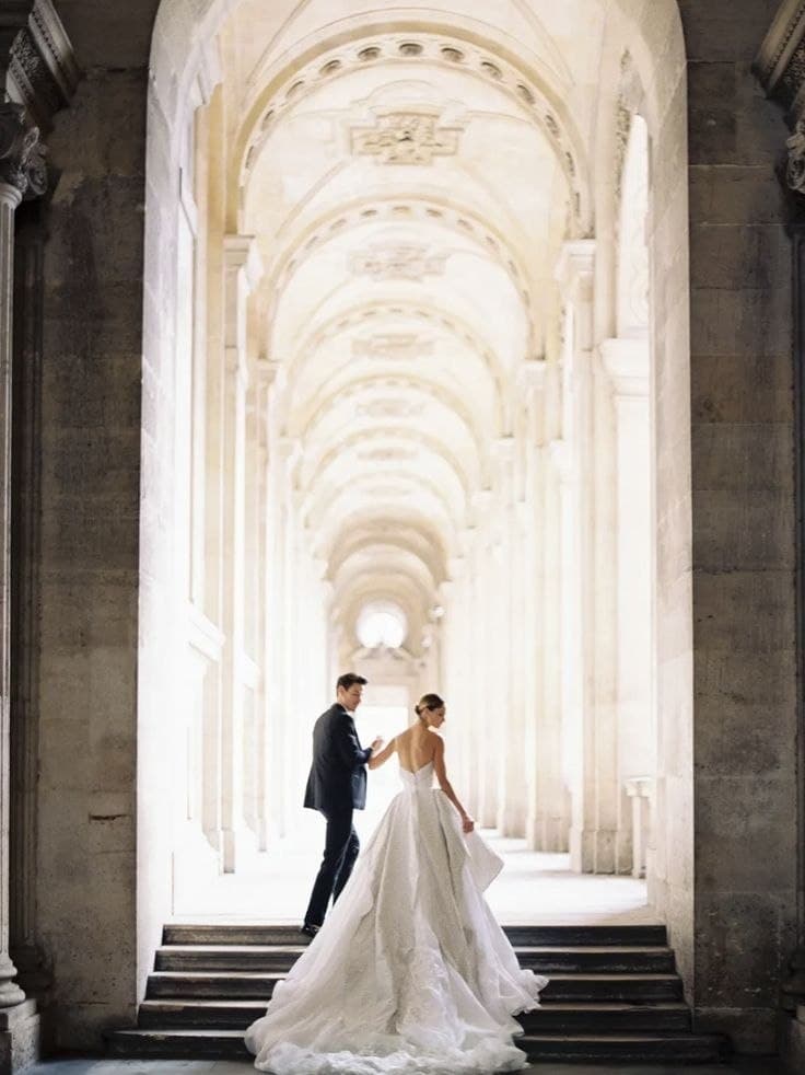 A couple in formal wedding attire beneath a grand sunlit colonnade